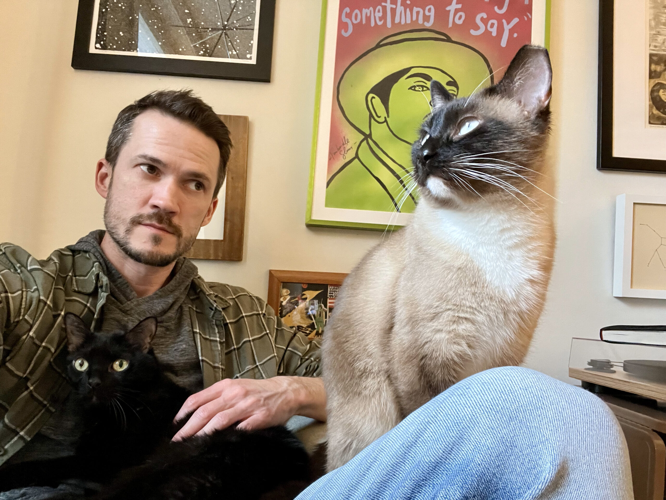 ID: Zach is a middle-aged man with brown hair and beard. He's sitting with two cats on his lap, one black, the other a siamese mix.